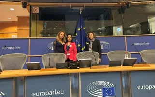 Mehrere Konferenzteilnehmerinnen stehen mit erhobenen Armen hinter den Tischen des Sitzungssaals.